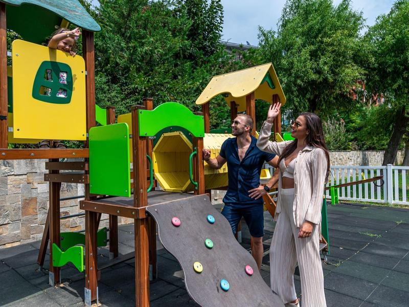 Zwei Personen stehen an einem bunten Spielplatzgerät aus Holz und Kunststoff im Freien.