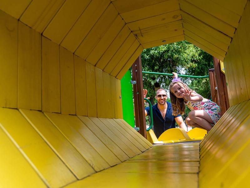 Blick durch eine gelbe Rutsche mit zwei Kindern, die lächelnd herausblicken im Freien.