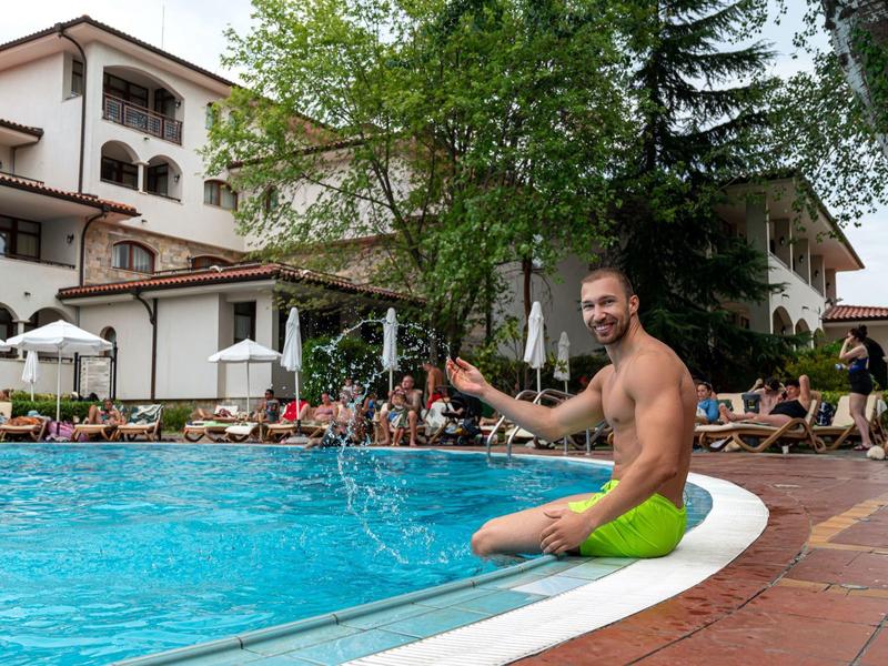 Mann in neongrüner Badehose sitzt am Rand eines blauen Pools, umgeben von Gästen und Gebäuden.