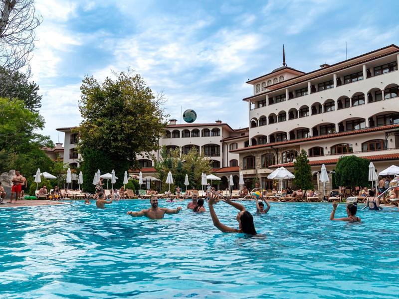Großer Pool mit mehreren Personen vor mehrstöckigem Hotel und blauem Himmel mit Wolken.