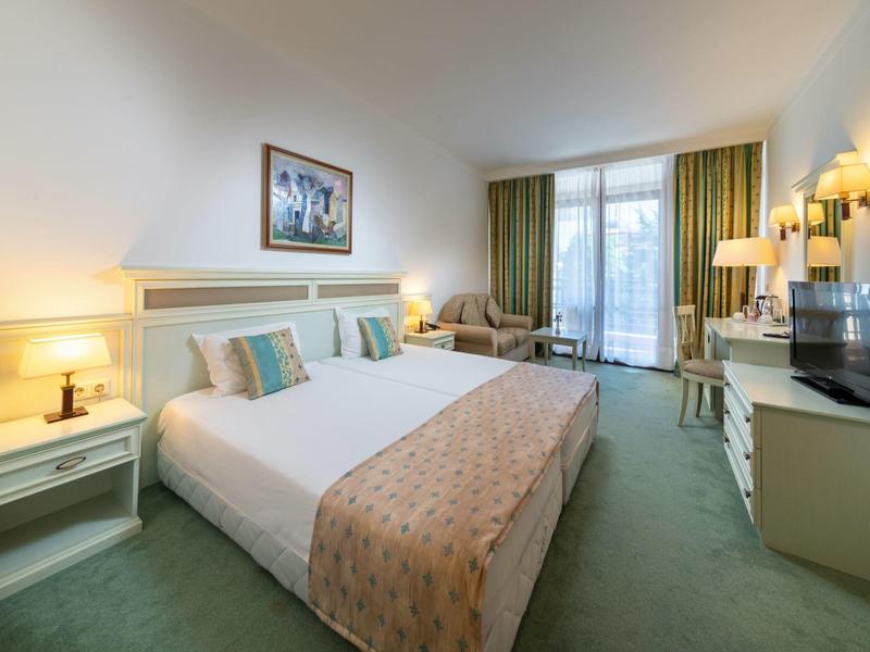 Helles Hotelzimmer mit großem Bett, Nachttischen, Sofa, TV und Balkon mit grünen Vorhängen.