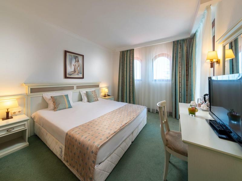 Hell eingerichtetes Hotelzimmer mit großem Bett, Fernseher, Schreibtisch und grünen Vorhängen.