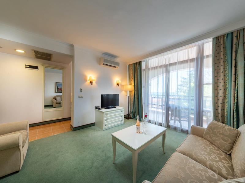 Helles Wohnzimmer mit grütem Teppich, beiger Couch, TV-Schrank und großem Fenster mit Vorhängen.