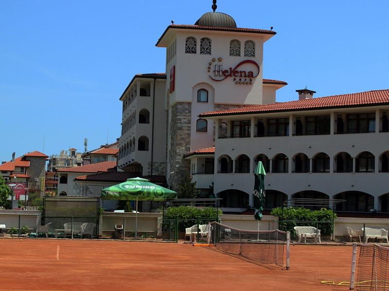 Großer Tennisplatz vor einem mehrstöckigen, weißen Gebäude mit roten Dächern und blauer Himmel.