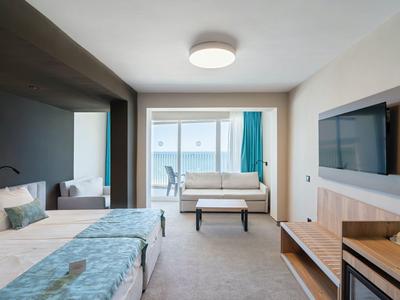 Modern hotel room with two beds, sofa, TV, and balcony view of the sea.