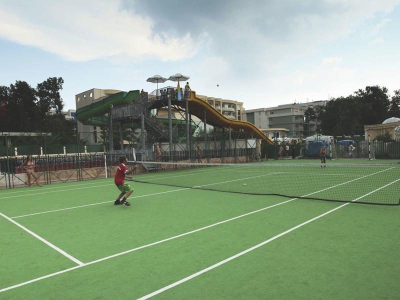 Tennis court with player in foreground and water slide in background under cloudy sky.