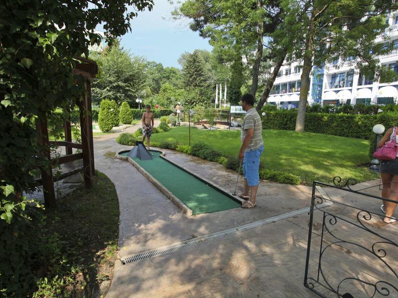 People playing mini golf in a green, shaded garden area next to a hotel.