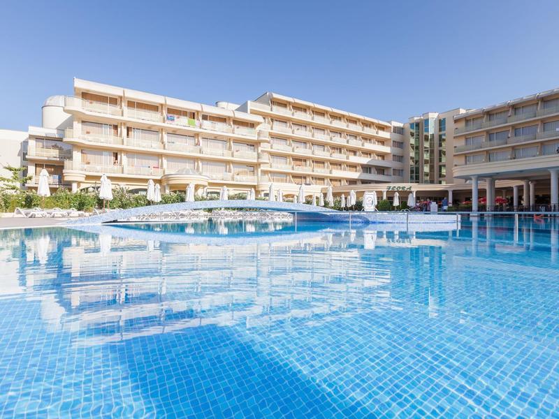 Large hotel building with a spacious outdoor pool and several sun umbrellas under clear sky.