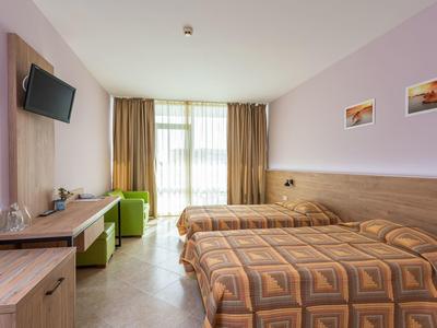 Helles Hotelzimmer mit zwei Einzelbetten, Holzmöbeln, grünem Sessel und großem Fenster mit Vorhängen.