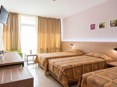Hell eingerichtetes Hotelzimmer mit drei Betten, Fernseher, Tisch und großem Fenster mit Vorhängen.