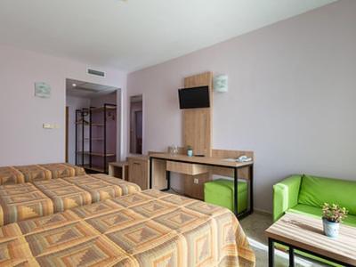 Hotelzimmer mit zwei Betten, grünem Sofa, Schreibtisch und Fernseher an heller Wand.