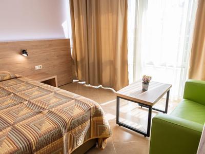Helles Schlafzimmer mit braunem Bett, Holzwand, grünem Sessel und kleinem Tisch am Fenster.