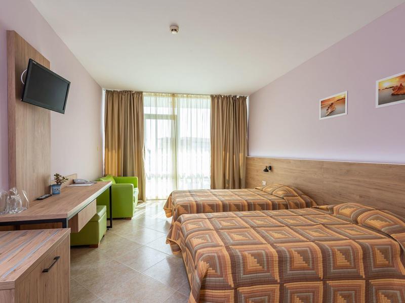 Hotelkamer met twee bedden, bureau, groene stoel en balkon met gordijnen.