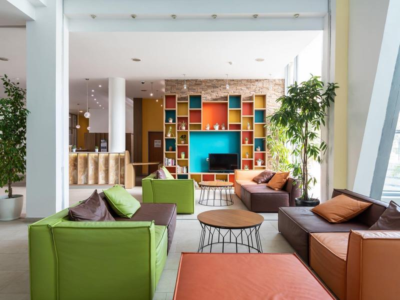 Heldere hotel lobby met kleurrijke banken, planten en een moderne boekenplank met tv.
