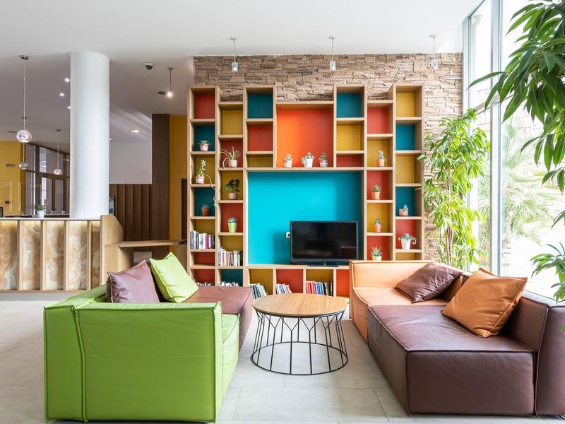 Lichte hotelkamer met kleurrijke planken, banken en groot raam met planten.