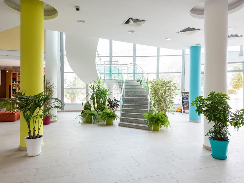 Helder verlichte hotellobby met grote ramen, trap en veel groene planten.
