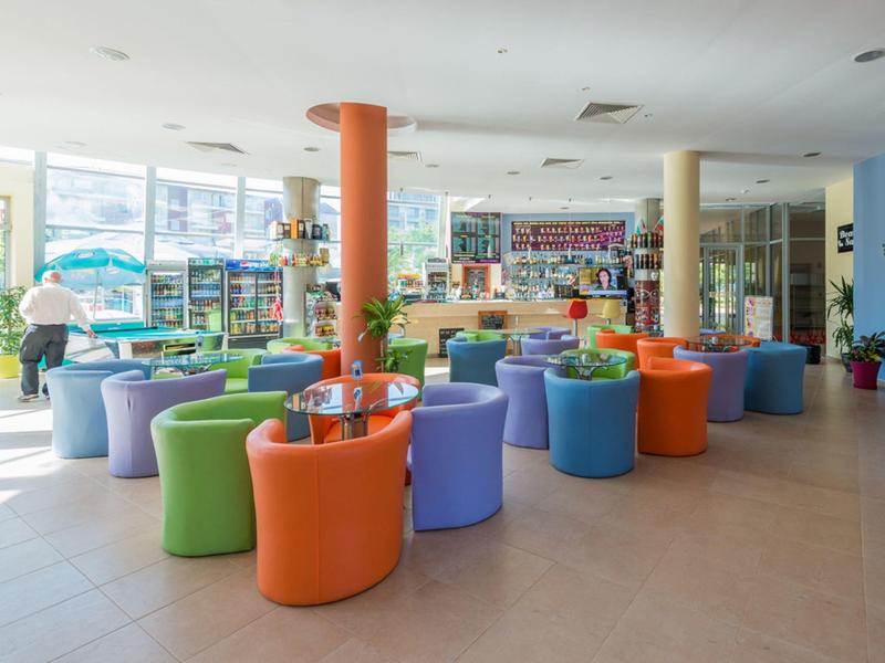 Lichte hotel lobby met kleurrijke stoelen en tafels, grote ramen en schappen met artikelen.
