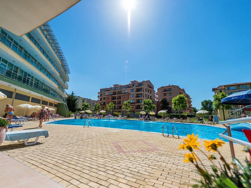 Groot buitenzwembad met ligstoelen, parasols en moderne hotelgebouwen onder een zonnige hemel.
