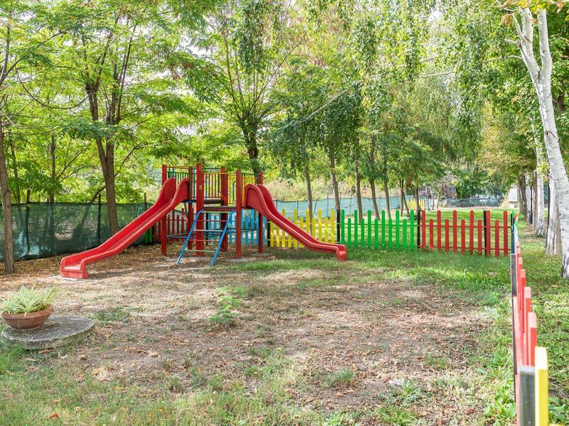 Speeltuin met rode glijbanen en kleurrijk hek in park omgeven door bomen.