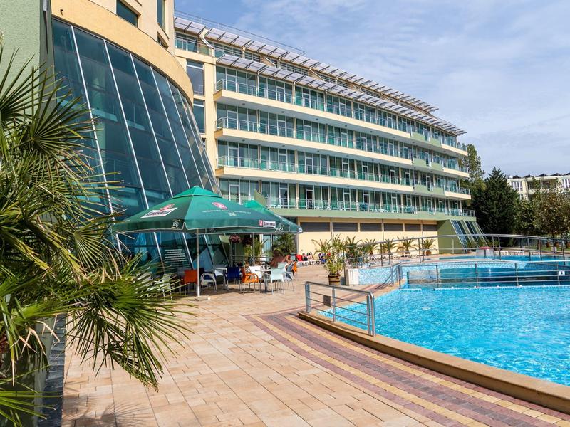 Modern hotelcomplex met zonneterras en groot buitenzwembad omgeven door palmbomen.