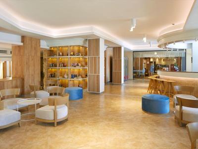 Großer, heller Raum mit Holzmöbeln, beigen Sesseln, blauen Poufs und indirekter Deckenbeleuchtung.