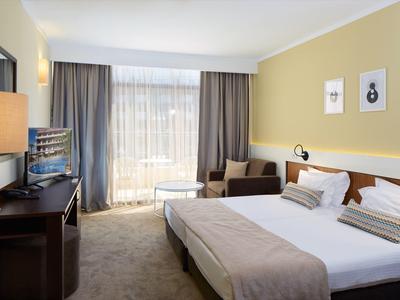 Hell eingerichtetes Hotelzimmer mit großem Bett, Sofa, TV und Fenster mit Vorhängen.