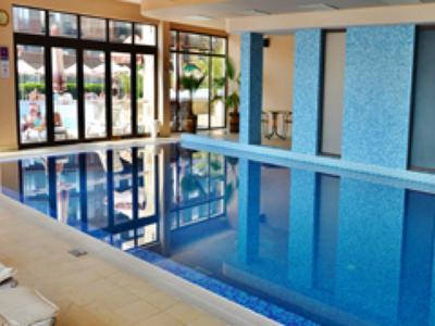 Innenraum mit rechteckigem Hallenbad, blauen Kacheln, großen Fenstern und Liegestühlen.