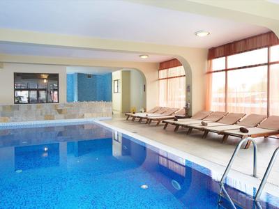 Innenbereich mit großem blauen Schwimmbad, Liegen und breiten Fenstern mit roten Vorhängen.