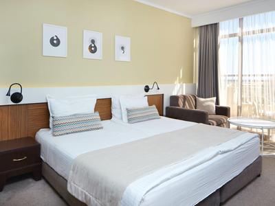 Modernes Hotelzimmer mit Doppelbett, Nachttisch, Sofasessel und großem Fenster mit Vorhängen.