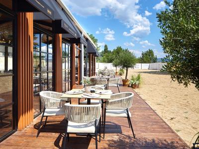 Terrasse mit weißen Stühlen und Tischen neben Sandfläche und Bäumen unter blauem Himmel.