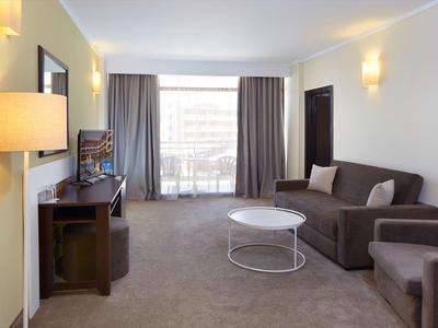 Helles Wohnzimmer mit grauem Teppich, Schlafsofa, Sessel, runden Couchtisch und großem Fenster mit Vorhängen.