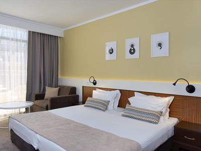 Helles modernes Schlafzimmer mit Doppelbett, grauen Vorhängen, Sessel und drei Bildern an gelber Wand.