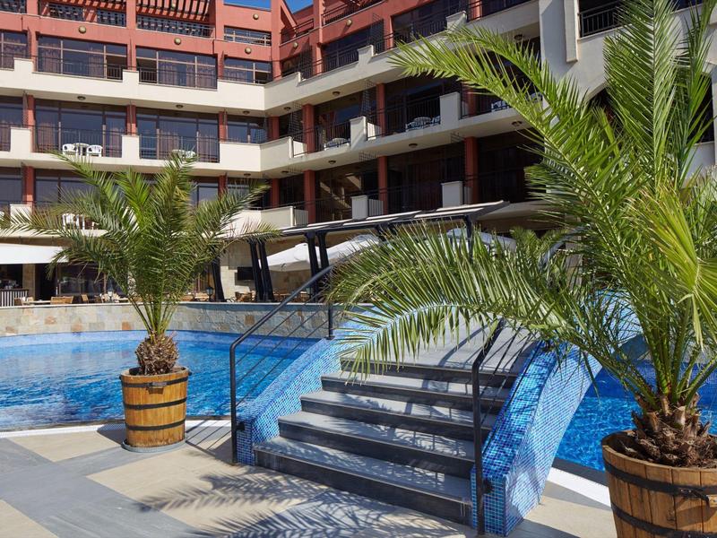 Hotelpool mit blauer Wasserfläche, Palmen in Holzfässern und mehrstöckigem Gebäude im Hintergrund.