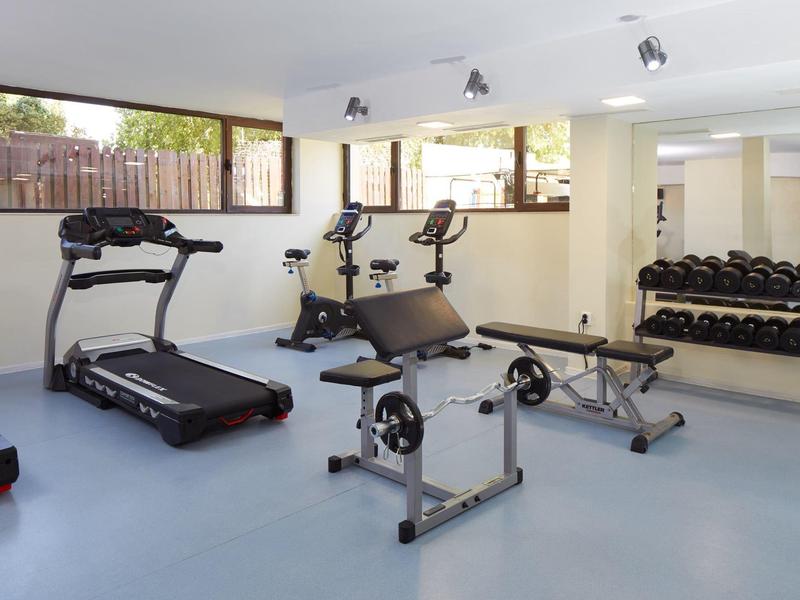 Heller Fitnessraum mit Laufband, Fahrrädern, Hantelbank, Langhantel und Spiegelwand.