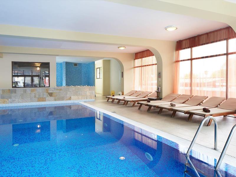 Innenbereich mit großem blauen Schwimmbad, Liegen und breiten Fenstern mit roten Vorhängen.