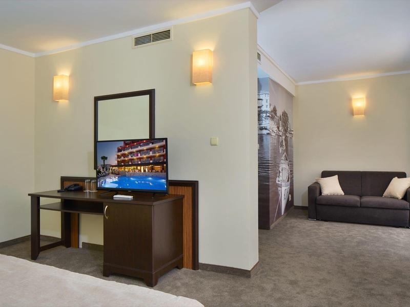 Helles Hotelzimmer mit Teppichboden, Schreibtisch, Fernseher, Sofa und moderne Wandlampen.