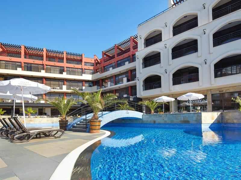 Moderner Hotelpool mit blauem Wasser, Liegestühlen, Sonnenschirmen und weißen Gebäuden im Hintergrund.