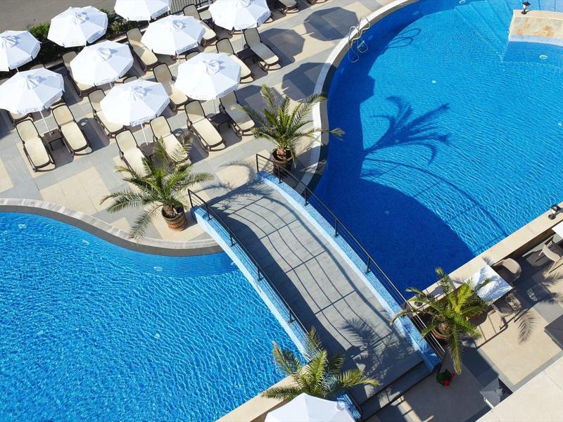 Blick auf zwei blaue Pools, eine Brücke dazwischen, Liegestühle und weiße Sonnenschirme.