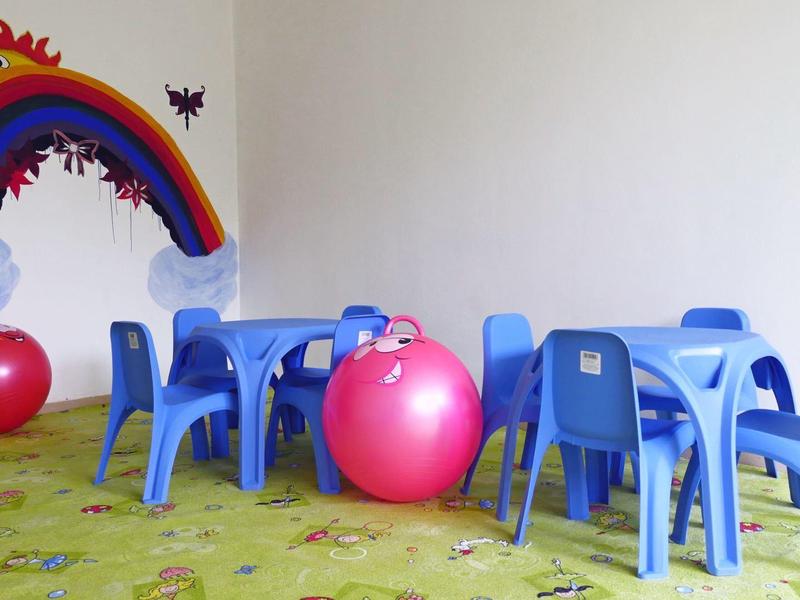 Kinderzimmer mit blauen Tischen und Stühlen, buntem Teppich und großer Wanddeko Regenbogen.