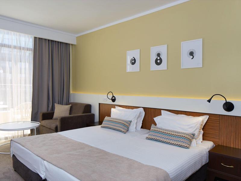 Helles modernes Schlafzimmer mit Doppelbett, grauen Vorhängen, Sessel und drei Bildern an gelber Wand.