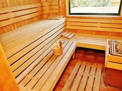 Sauna de madera con banco, cubo y cucharón, y una ventana que deja entrar la luz del día.