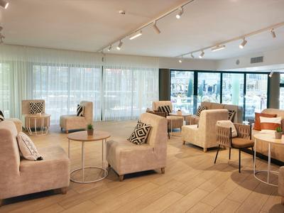 Área de salón luminosa en hotel con sillones beige, sofás y mesas pequeñas sobre suelo de madera.