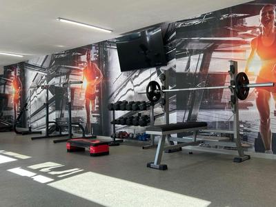 Moderner Fitnessraum mit Gewichten und Trainingsgeräten vor einer großen Wand mit Sportler-Silhouetten.