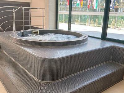 Jacuzzi gris moderno con escalones y pasamanos en una habitación luminosa junto a las ventanas.