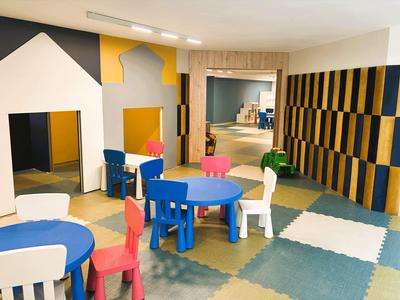 Sala de juegos infantil colorida con mesas, sillas y casitas sobre piso acolchonado.