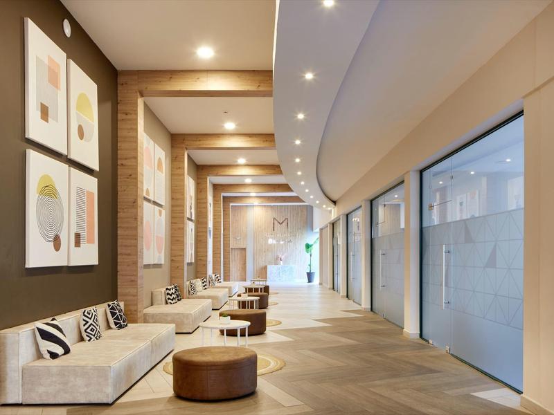 Pasillo moderno y luminoso de hotel con bancos, puf redondo y arte abstracto en la pared.