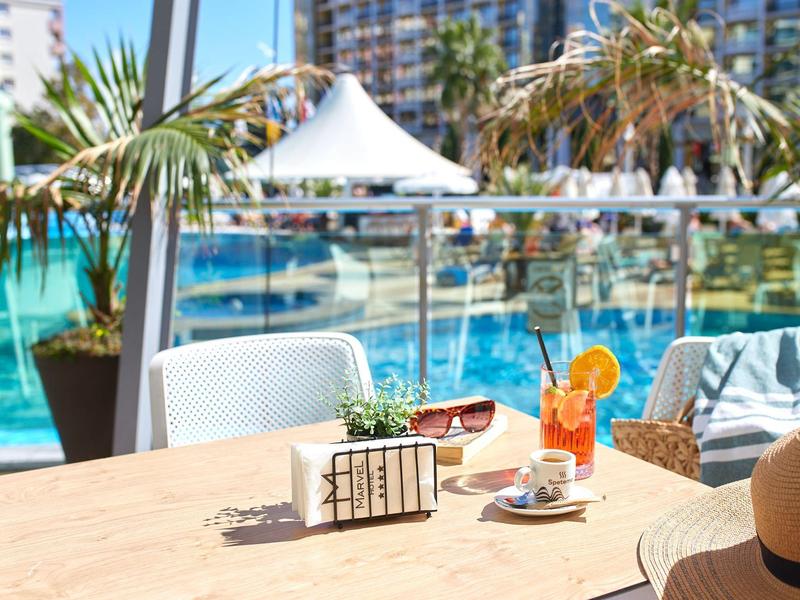 Una mesa en una terraza de hotel con bebidas y hojas de palma, piscina y edificio del hotel al fondo.