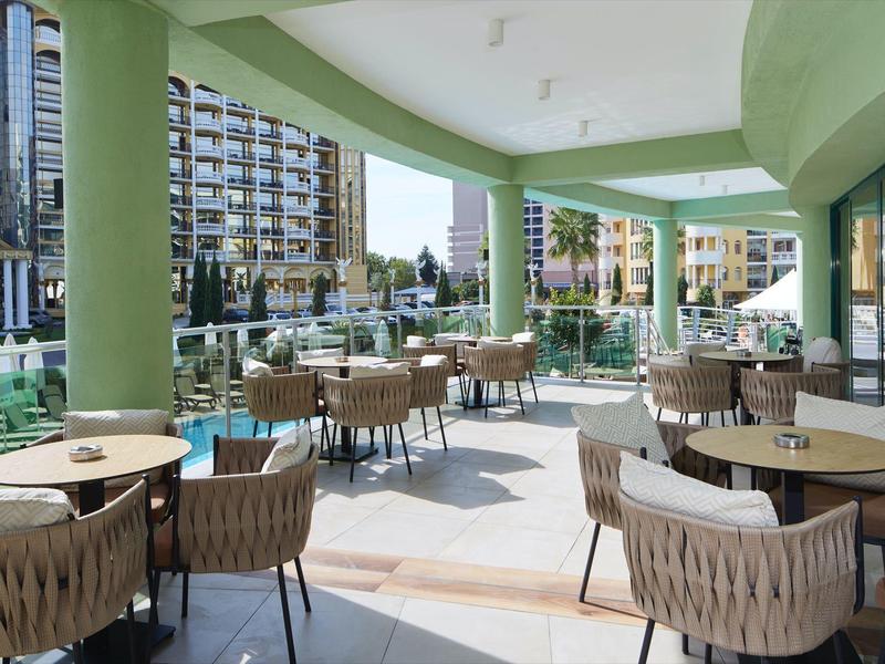 Terraza cubierta de hotel con mesas y sillas, rodeada de hoteles y palmeras.