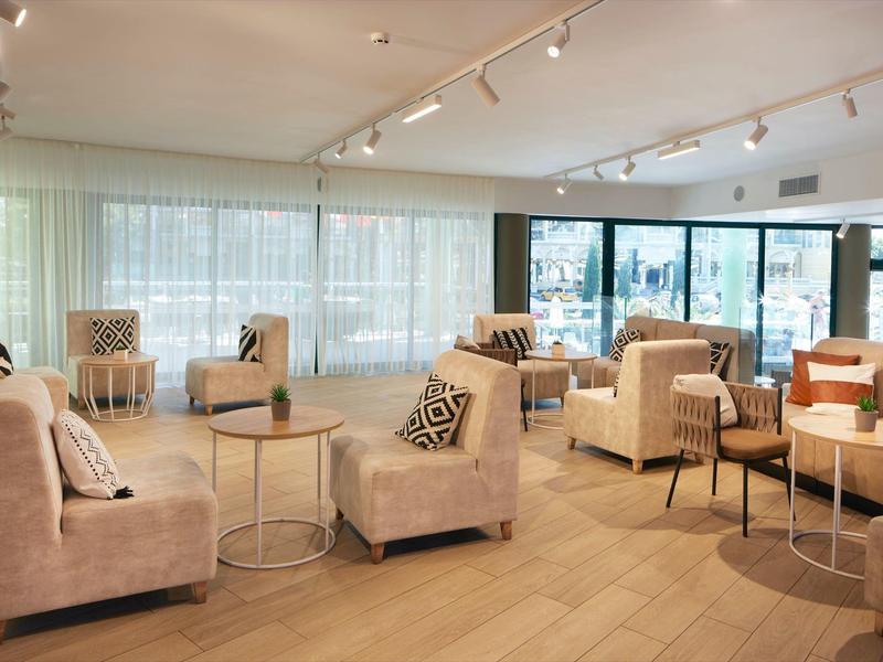 Área de salón luminosa en hotel con sillones beige, sofás y mesas pequeñas sobre suelo de madera.