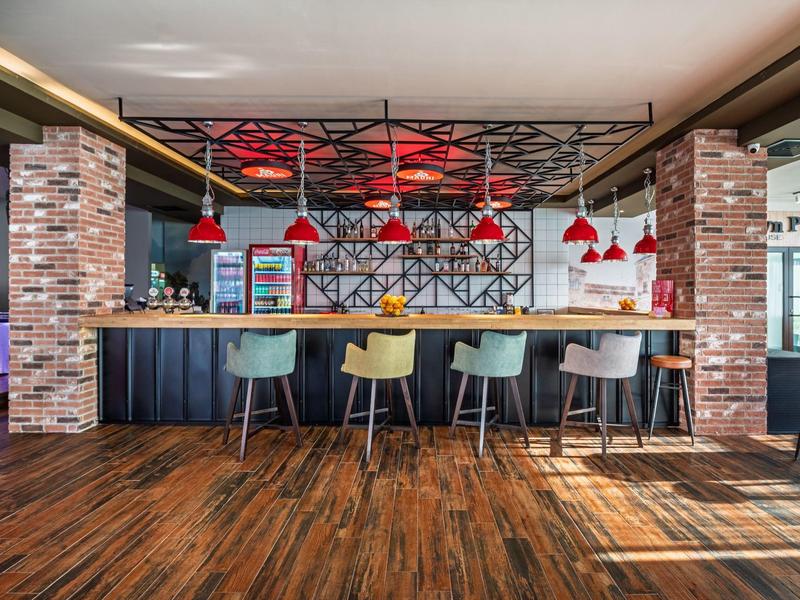 Área de bar moderna en hotel con suelo de madera, taburetes coloridos e iluminación roja